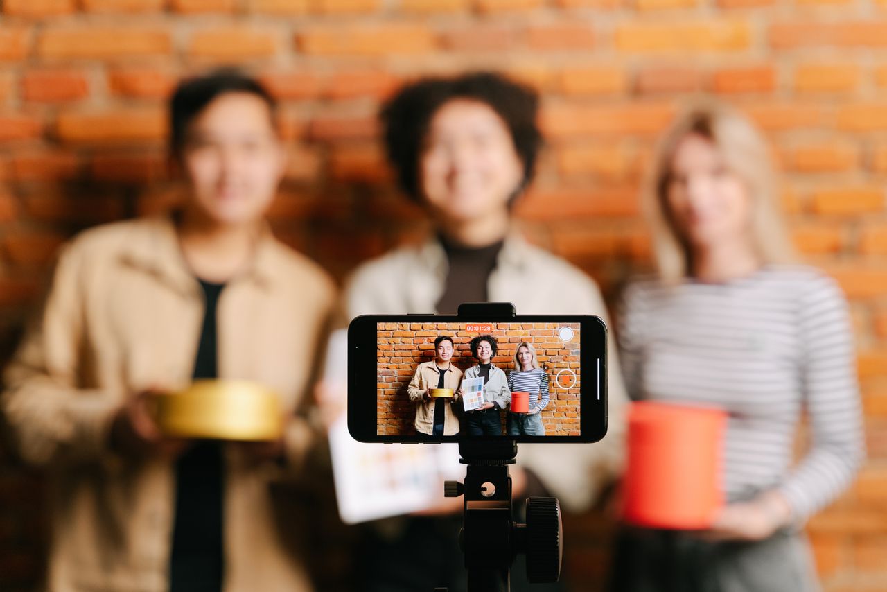 A varied group of vloggers filming a video with a smartphone in front of a brick wall background.